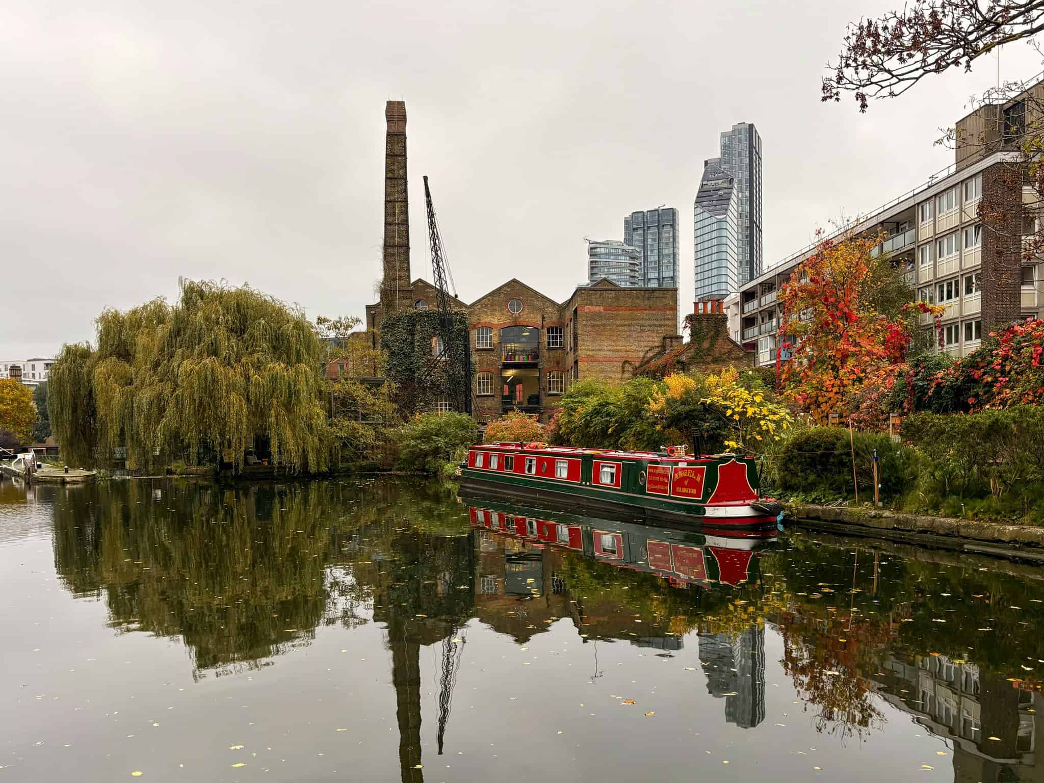 Charming canal scene with colorful houseboat, lush autumn foliage, modern high-rise buildings, historic brick factory, and overcast sky in London.