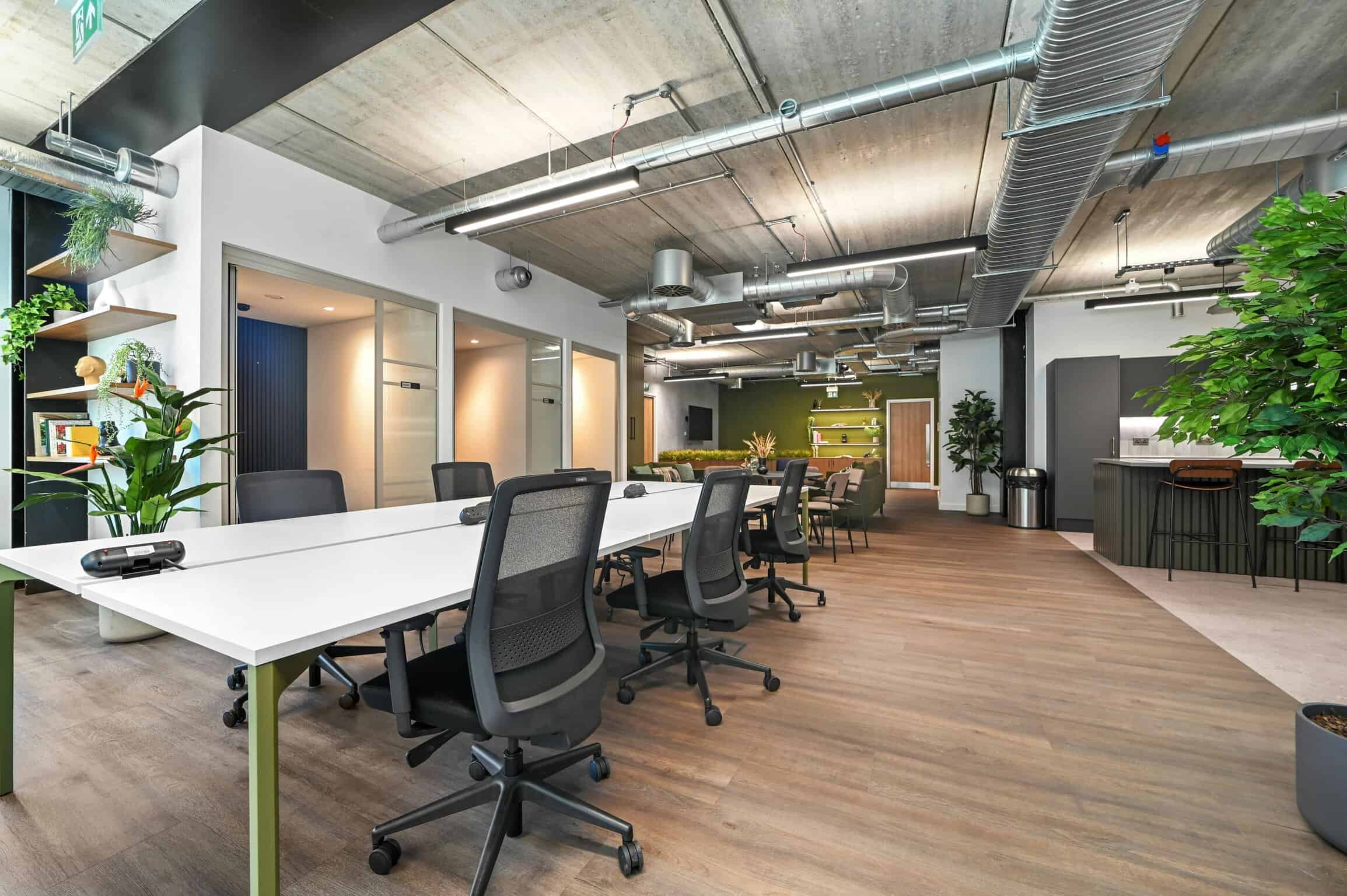 Modern coworking office space with open-plan desks, ergonomic chairs, and industrial-style ceilings featuring exposed ducts and piping, surrounded by greenery and contemporary decor.