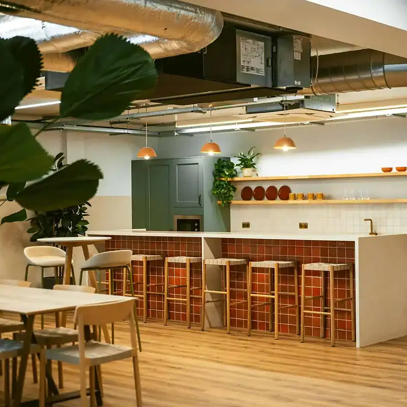 Contemporary office kitchen in Shoreditch with green cabinets, wooden bar stools, and open ceiling with ductwork, ideal for coworking spaces and professional environments.