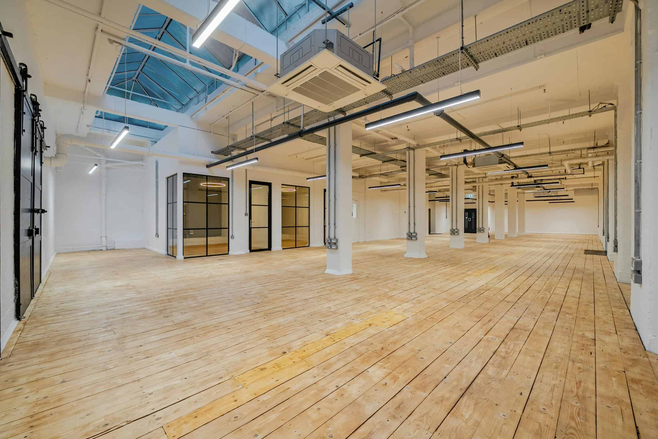 A spacious, modern industrial-style interior with high ceilings, large windows, exposed ductwork, and wooden flooring, ideal for office or creative workspace use.