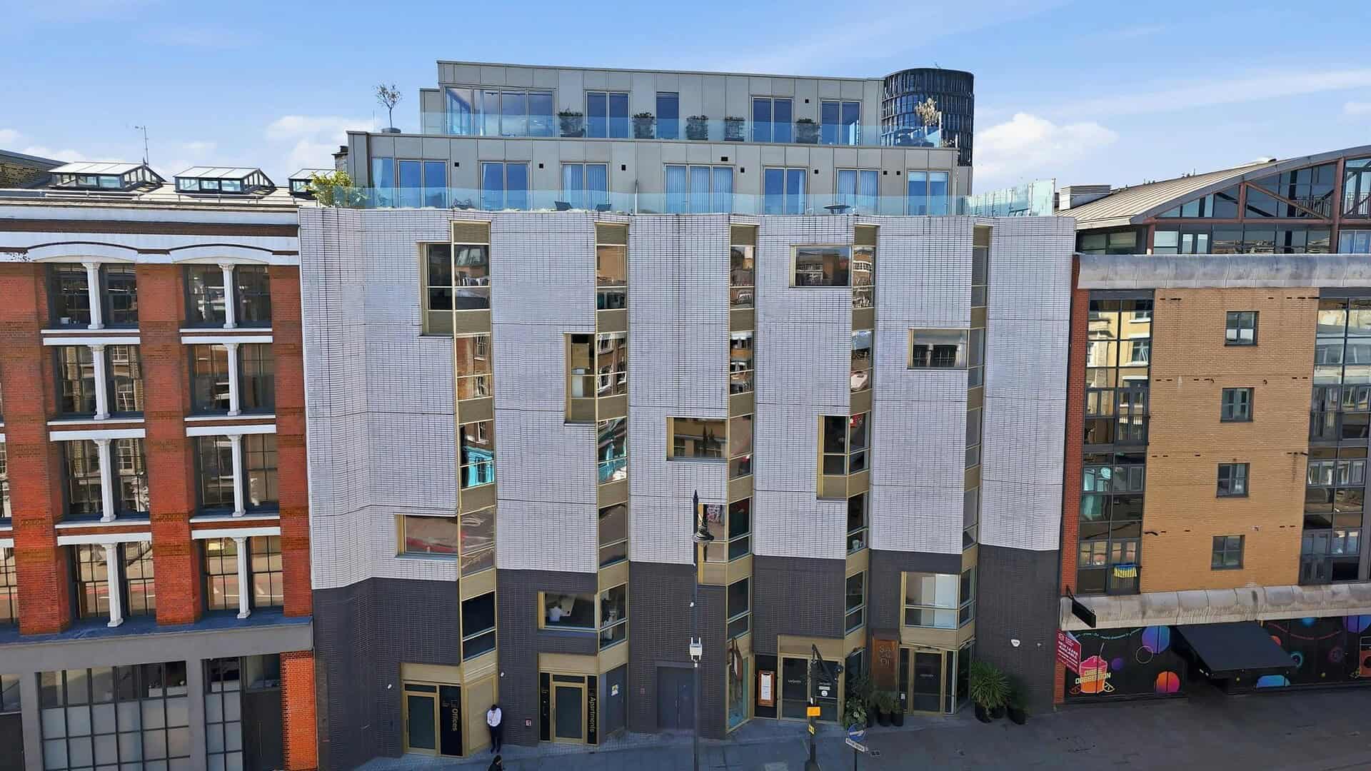 Modern multi-story residential building with a mix of glass and metal facade, situated in an urban area with neighboring structures, under a clear blue sky.