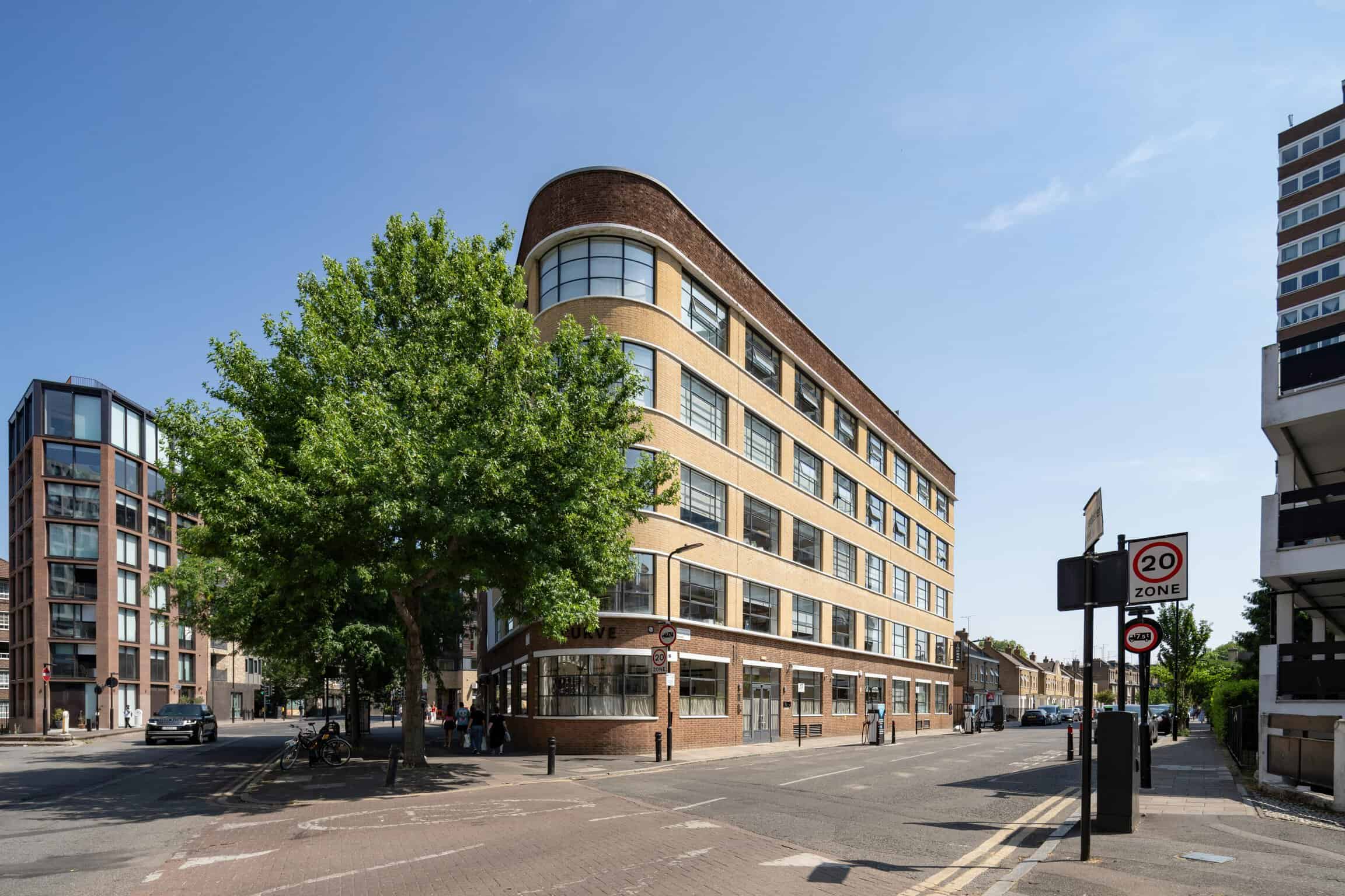 Modern multi-story building at the corner of Haberdasher Street with large windows and rounded corner design, surrounded by trees and urban street scene in London.