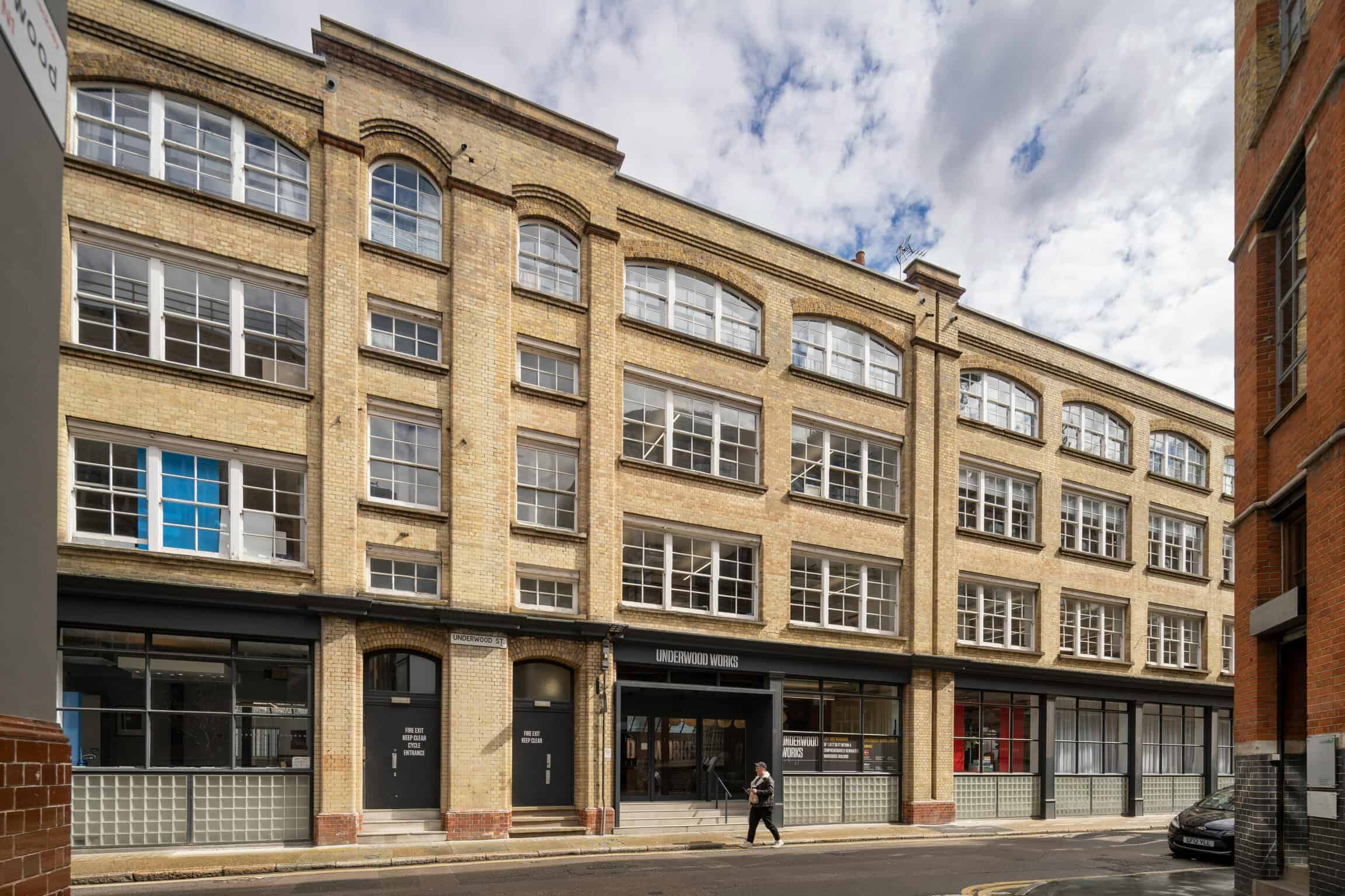 A modern mixed-use building with large windows and brick facade in an urban setting, featuring retail spaces on the ground floor and office or residential units above, under a partly cloudy sky.