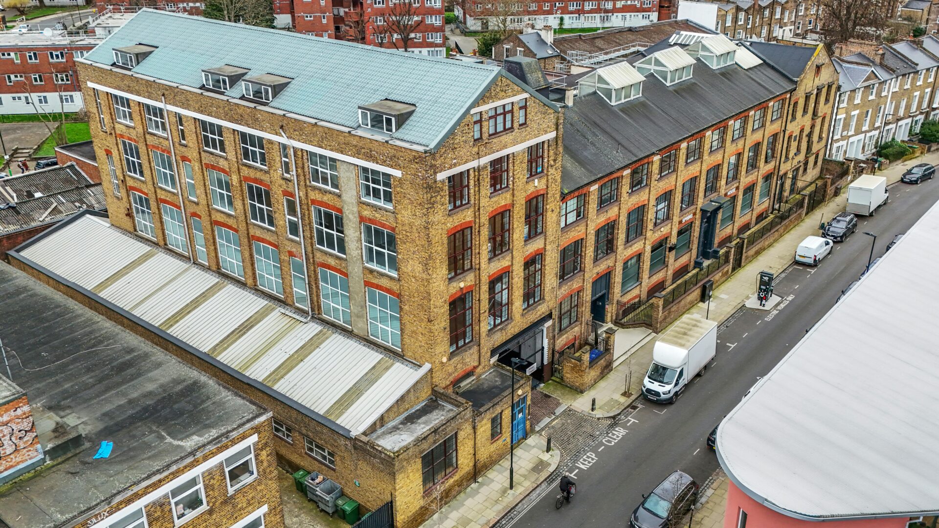 A modern brick building with large windows and a light blue roof, situated along a city street with parked cars and pedestrians, showcasing contemporary commercial or office space in London.