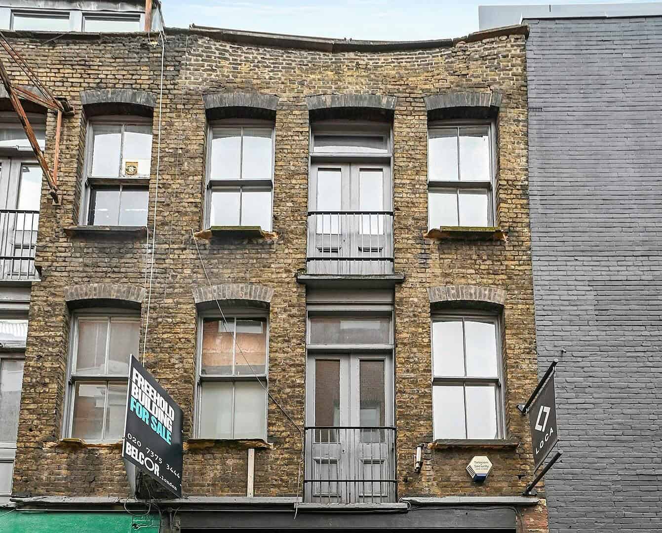 A modern commercial building with a brick facade, large glass windows with decorative metal grilles, and a black entrance door, situated between a green storefront and a gray building in an urban sett.