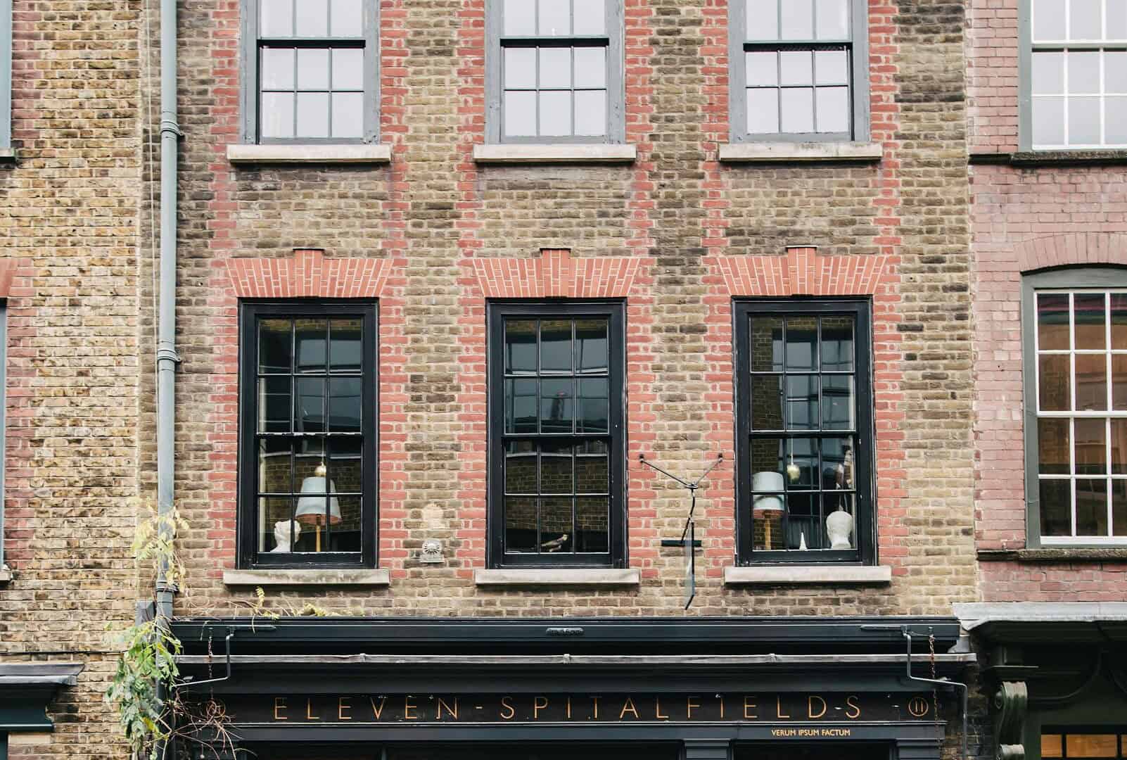 A modern brick building with large black-framed windows and a black storefront labeled "Eleven Spitalfields," located on a city street with parked cars and greenery.