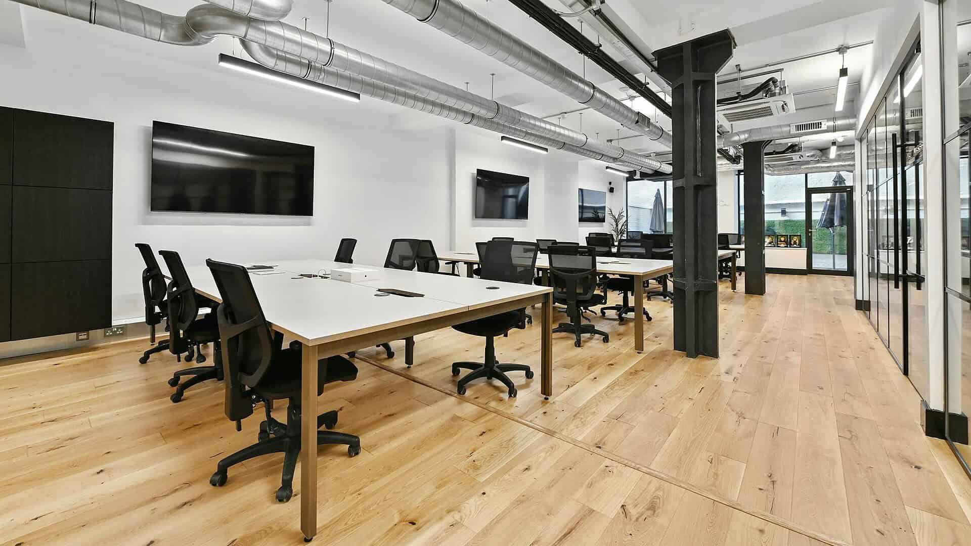 Flexible modern office space with large meeting tables, ergonomic chairs, and multiple wall-mounted screens in a bright, open-plan environment with industrial design elements.
