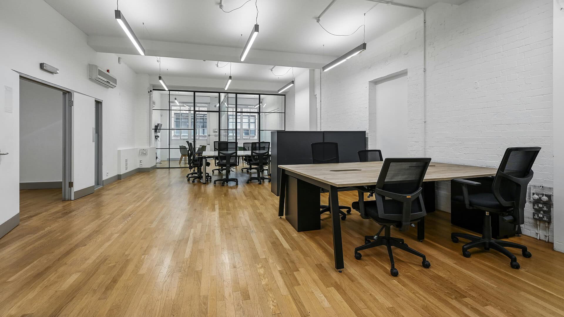 Modern office space with wooden flooring, white walls, large windows, and contemporary furniture including desks and chairs, ideal for professional work environments.