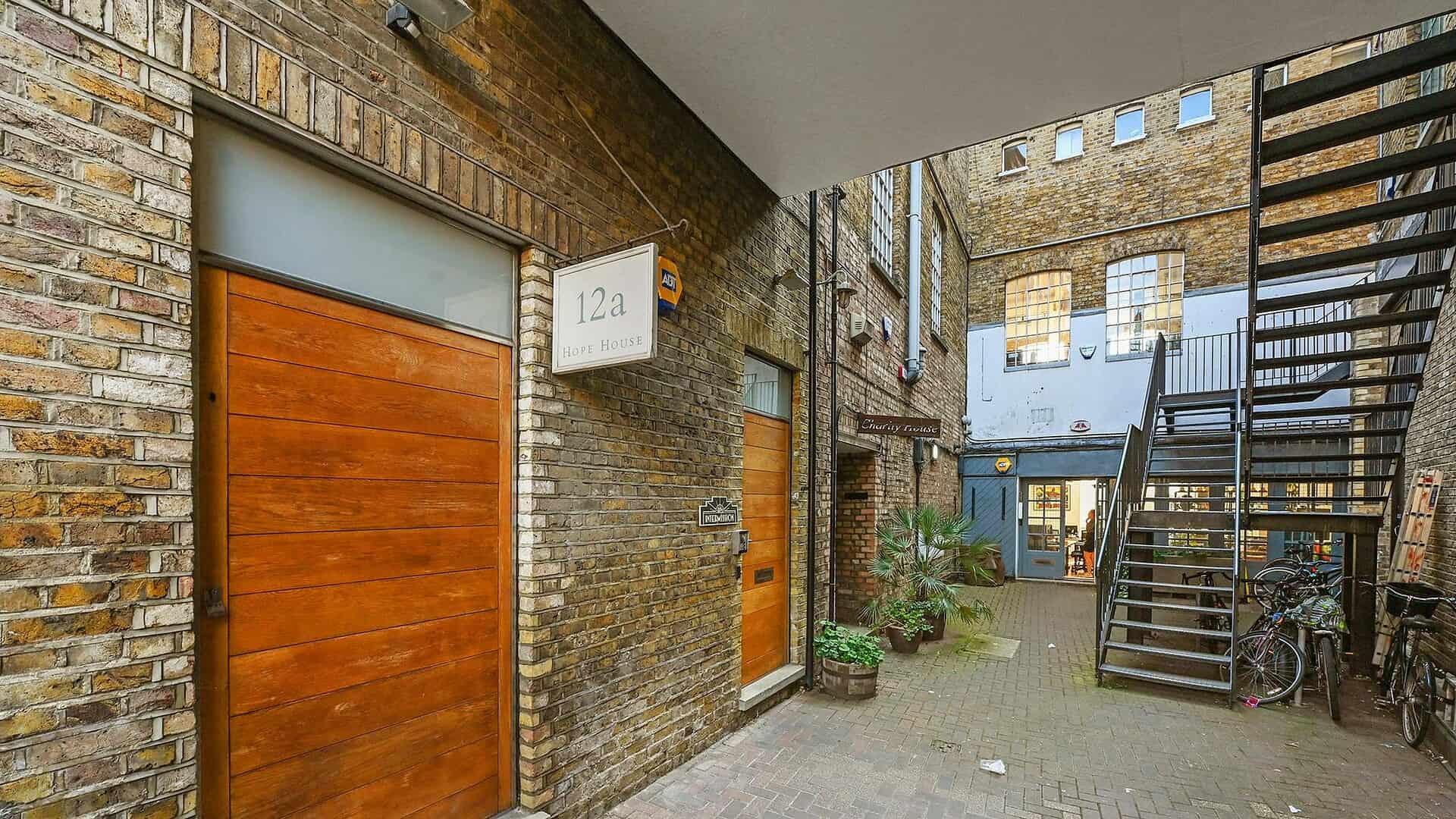 Courtyard view of Hope House Perseverance Works in East London, featuring brick walls, wooden doors, outdoor stairs, and bicycles, highlighting community and urban resilience.
