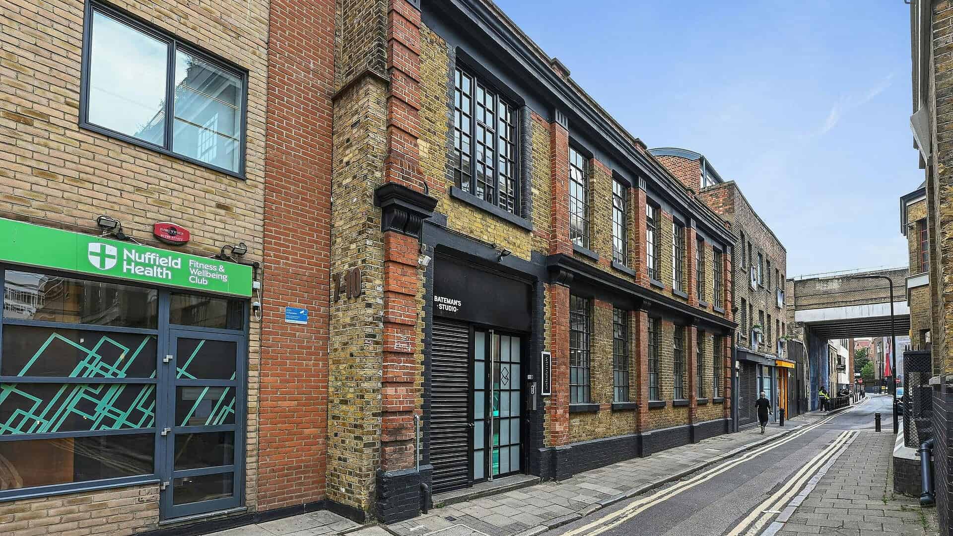 Modern office building in London with brick facade, large windows, and black accents, located on a narrow street with a nearby health clinic and pedestrian walkway.