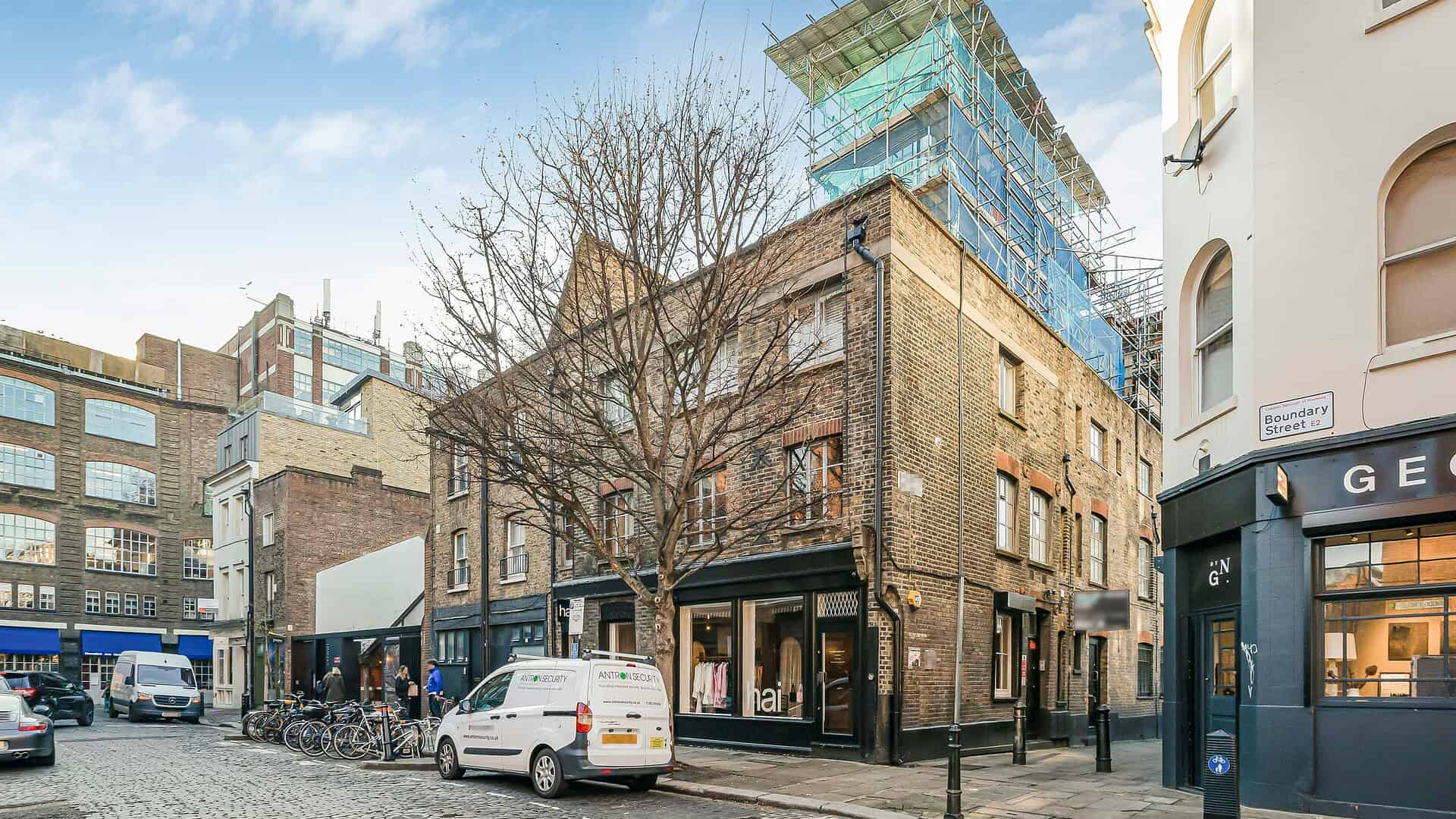 Urban street scene featuring a historic brick building at Boundary Passage with modern construction scaffolding, parked cars, and surrounding shops in London.