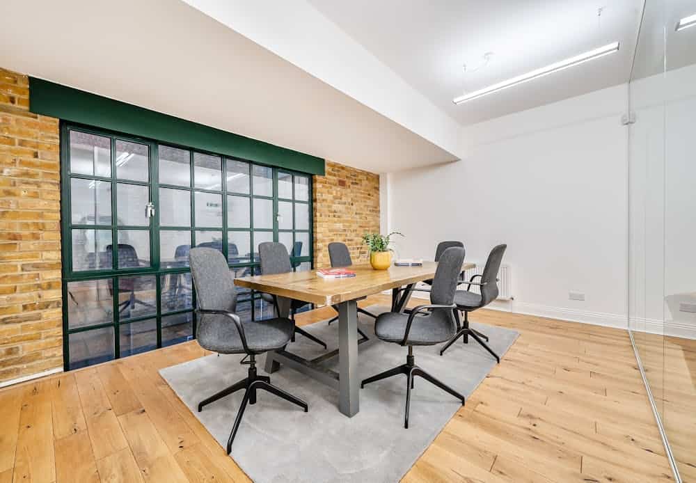 Contemporary office meeting room with a wooden table, six chairs, exposed brick wall, large window with green frame, and minimalist decor in Shoreditch.