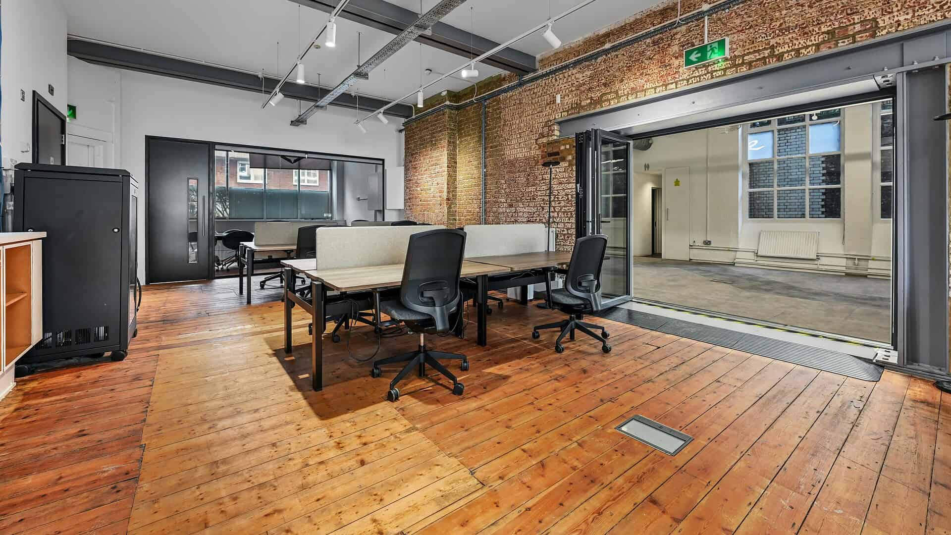 Spacious modern office with wooden floors, exposed brick walls, large windows, and flexible workstations in a stylish London building.