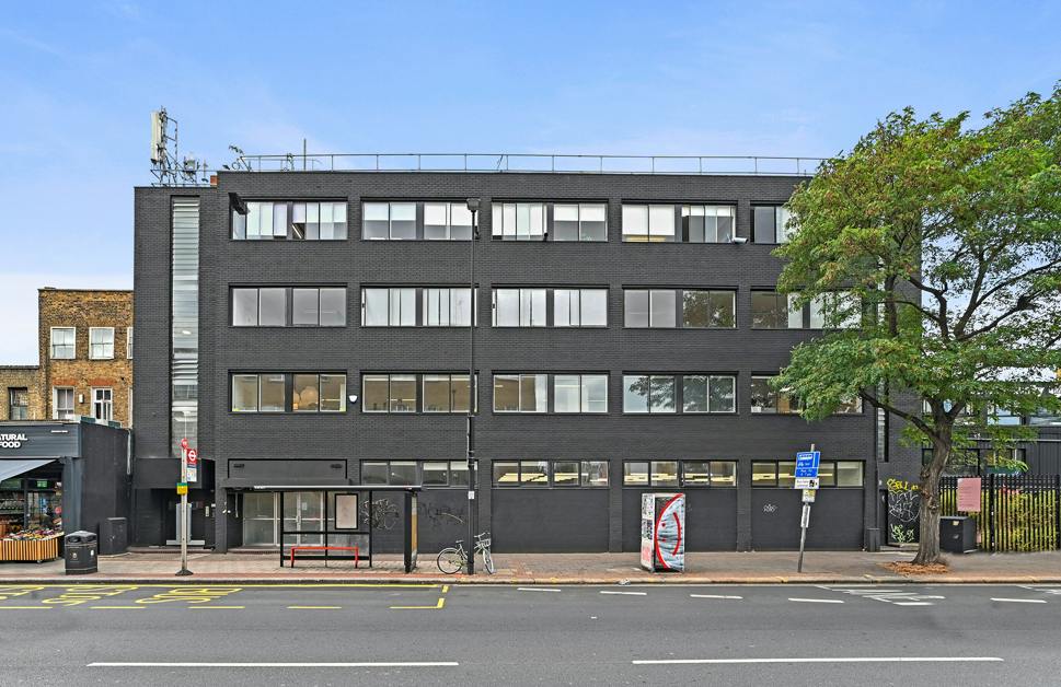 Contemporary black office building on Hackney Road with large windows, situated next to a tree and street furniture, under a clear blue sky.