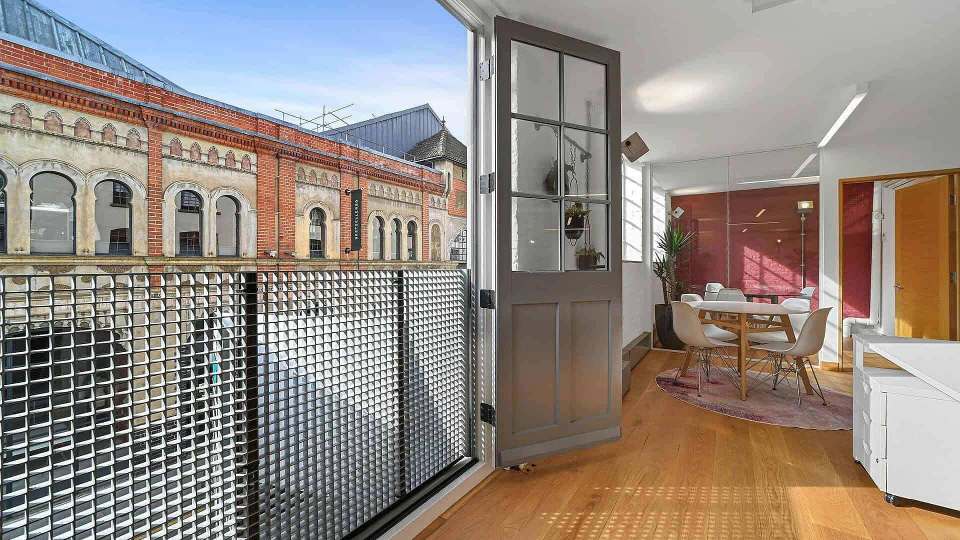 A modern interior living space with a balcony overlooking historic brick buildings, featuring wooden flooring, contemporary furniture, and large windows allowing natural light.