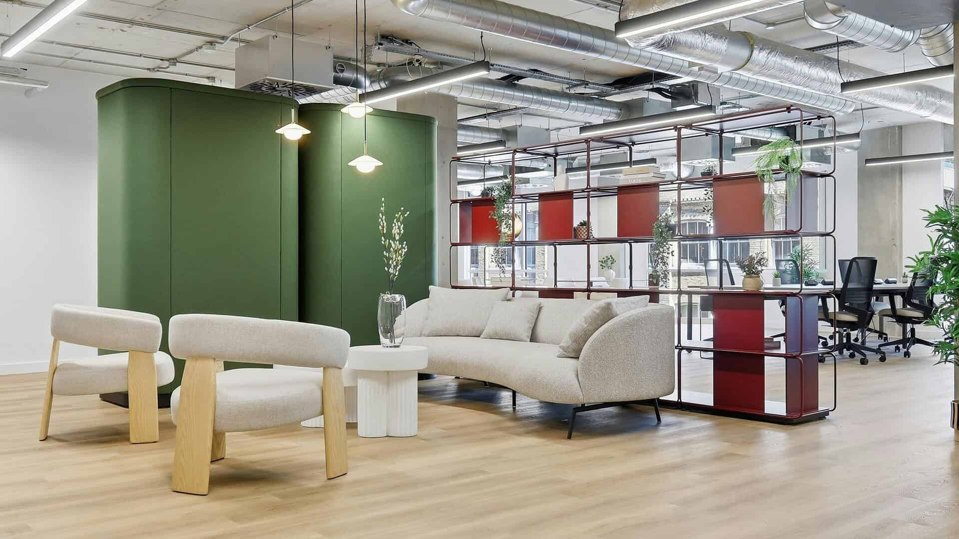 Modern office lounge area with beige seating, green accent wall, and open-plan workspace with red and glass partition, natural light, and contemporary decor for collaborative work environments.