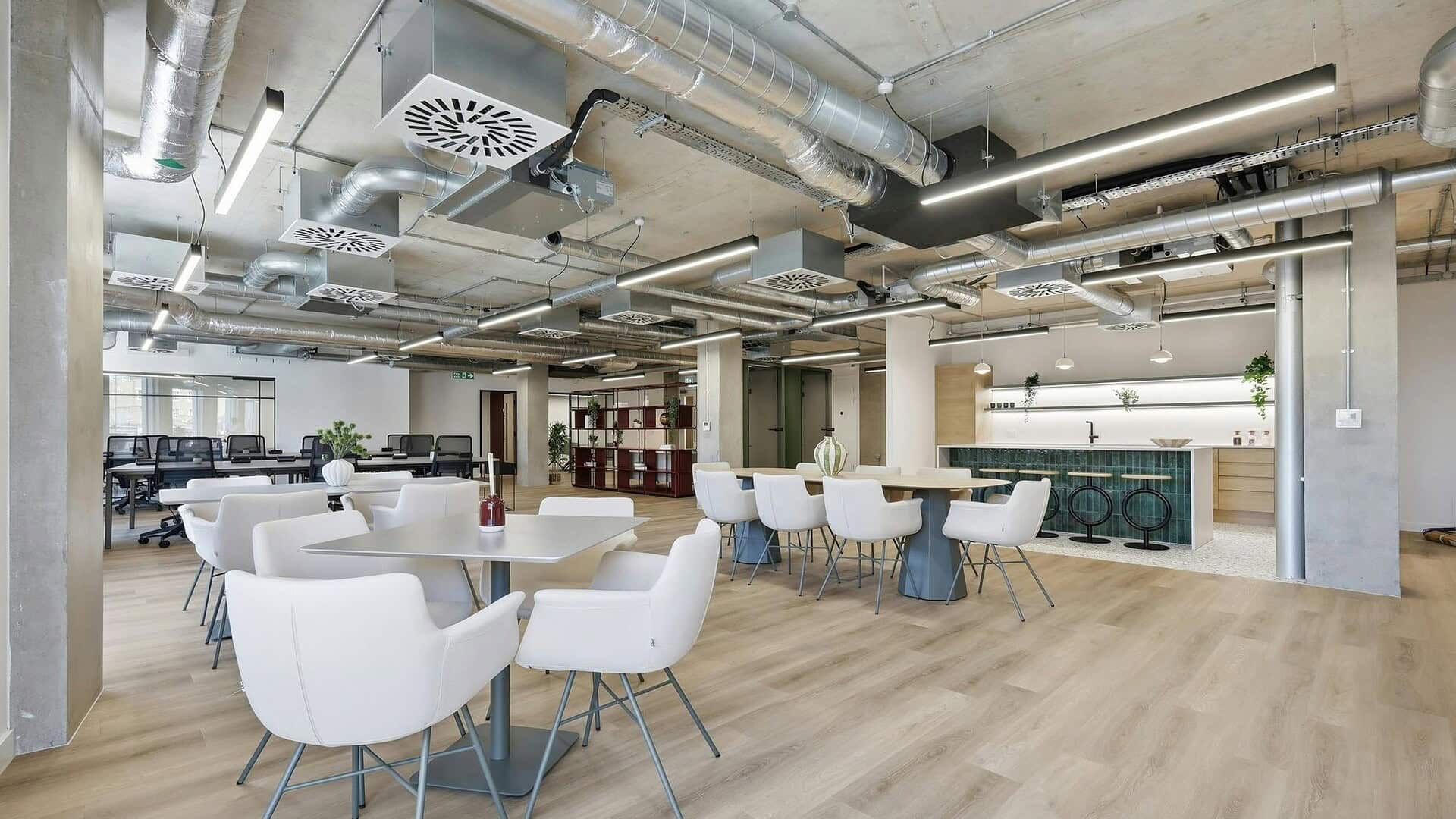 Modern office lounge area with white chairs, wooden flooring, exposed ceiling pipes, and contemporary decor, ideal for collaborative work and relaxation.