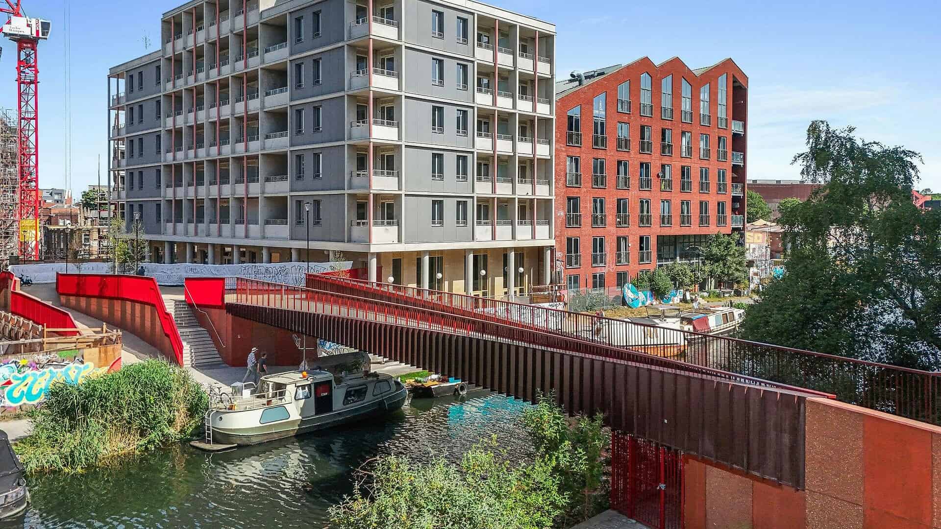 Contemporary residential buildings along the river with a pedestrian bridge, boats, and lush greenery in an urban setting.