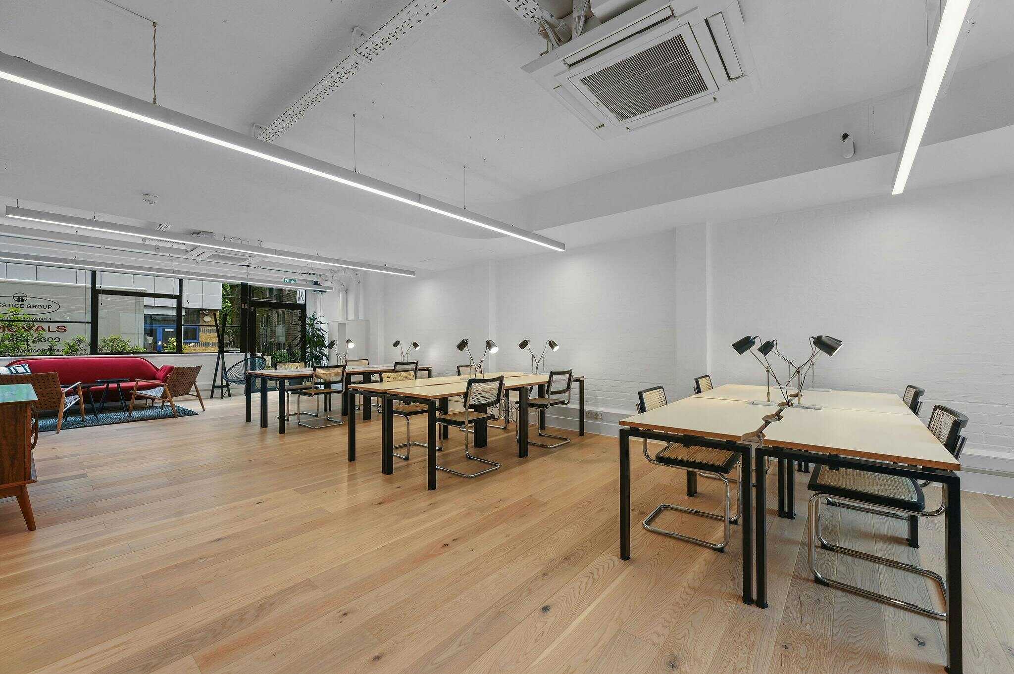 A modern, minimalist coworking space with white walls, wooden flooring, and contemporary furniture, featuring adjustable desk lamps and large windows for natural light.