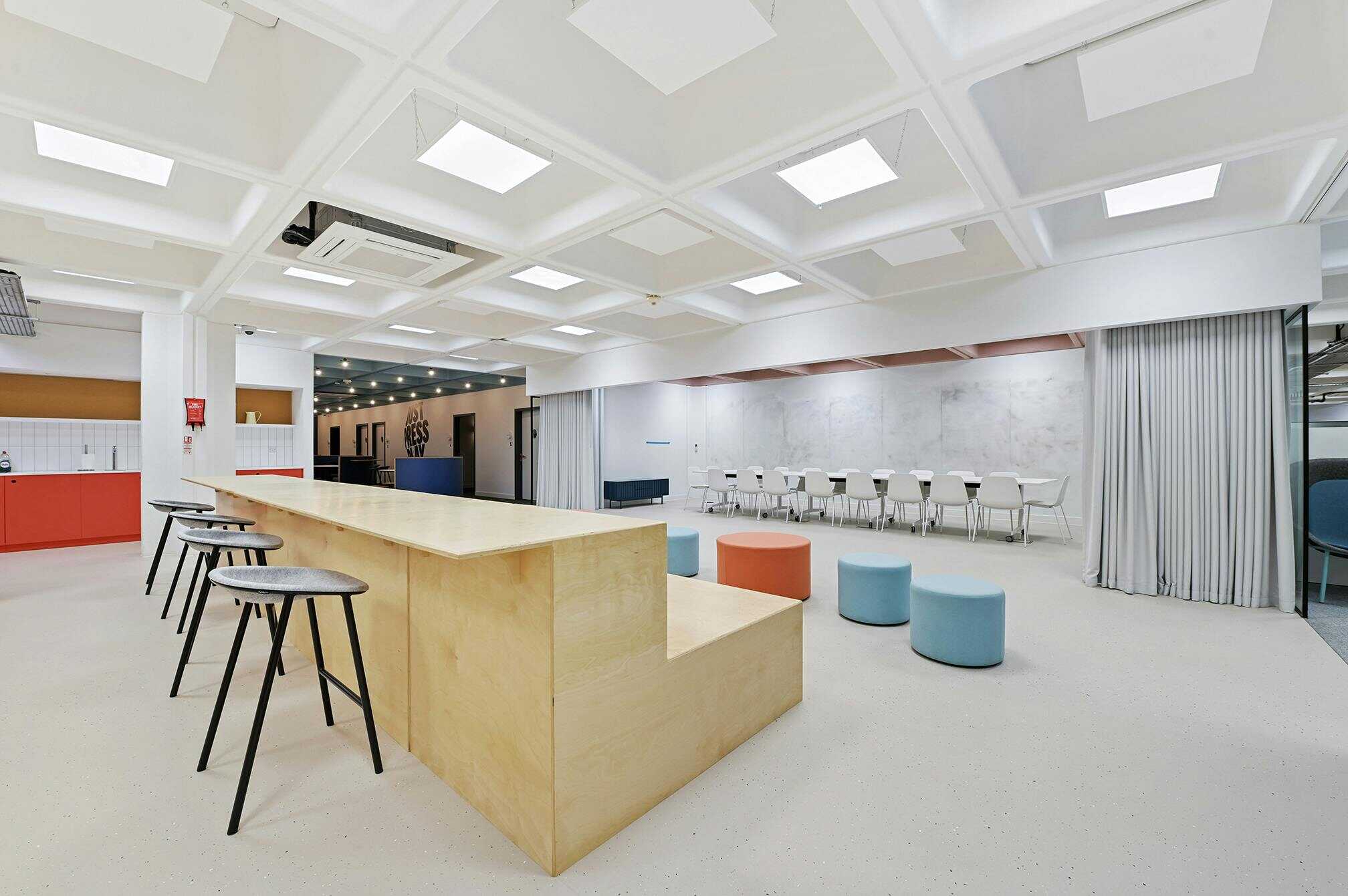 Modern, bright office lounge with a large wooden table, bar stools, colorful ottomans, and a row of chairs along the wall, featuring a sleek, contemporary design and ample natural lighting.