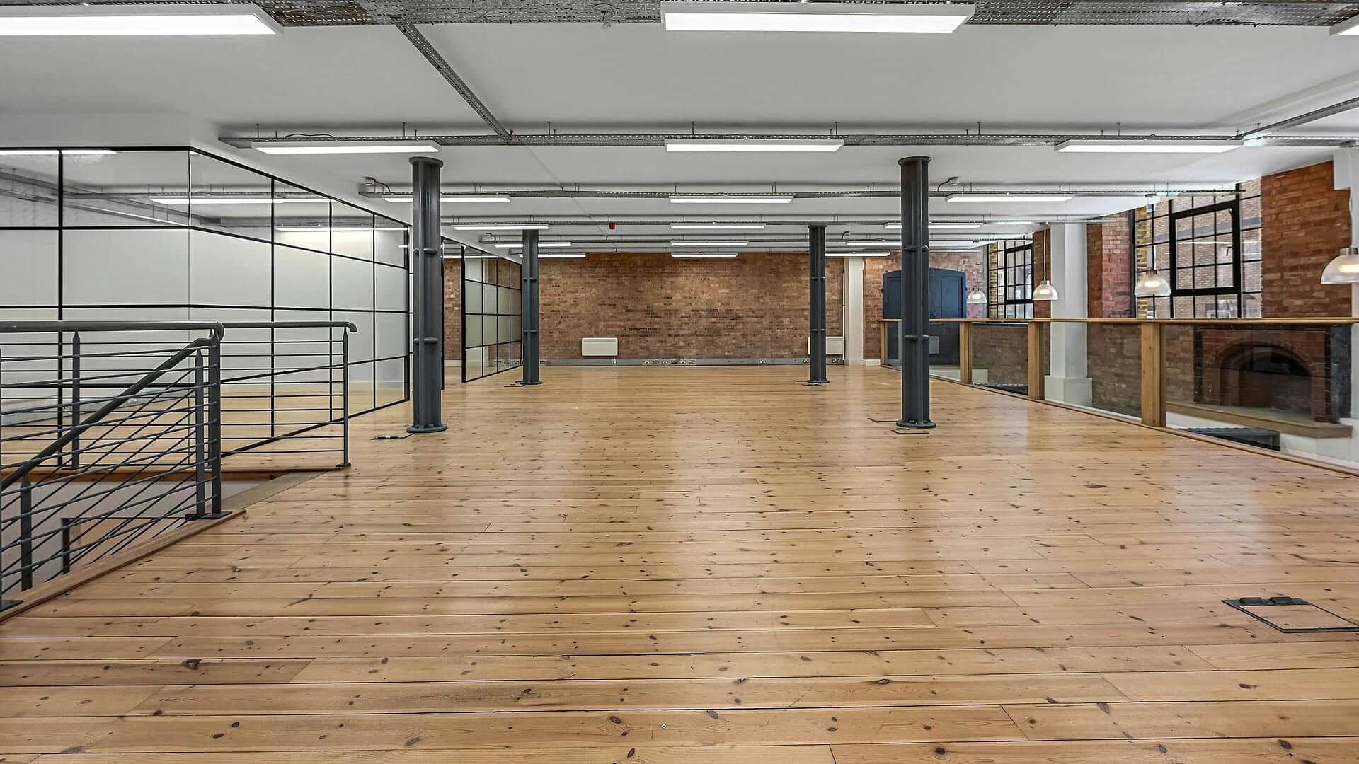 A spacious, modern industrial-style interior with polished wooden floors, exposed brick walls, large windows, and metal support columns, ideal for creative workspaces or events.
